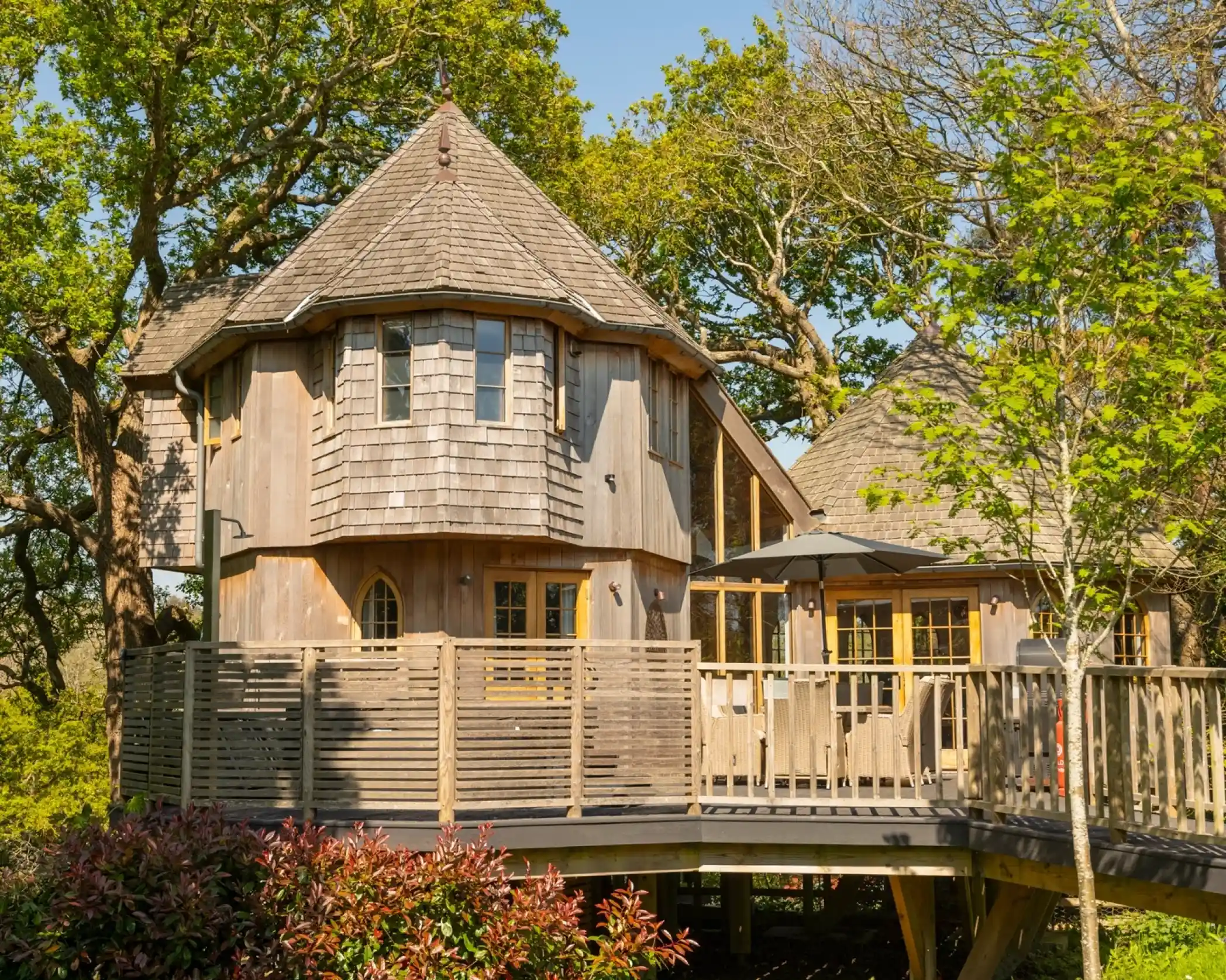 A unique wooden building with a circular design, featuring a shingled roof and large windows. It is elevated on a deck surrounded by greenery and trees.
