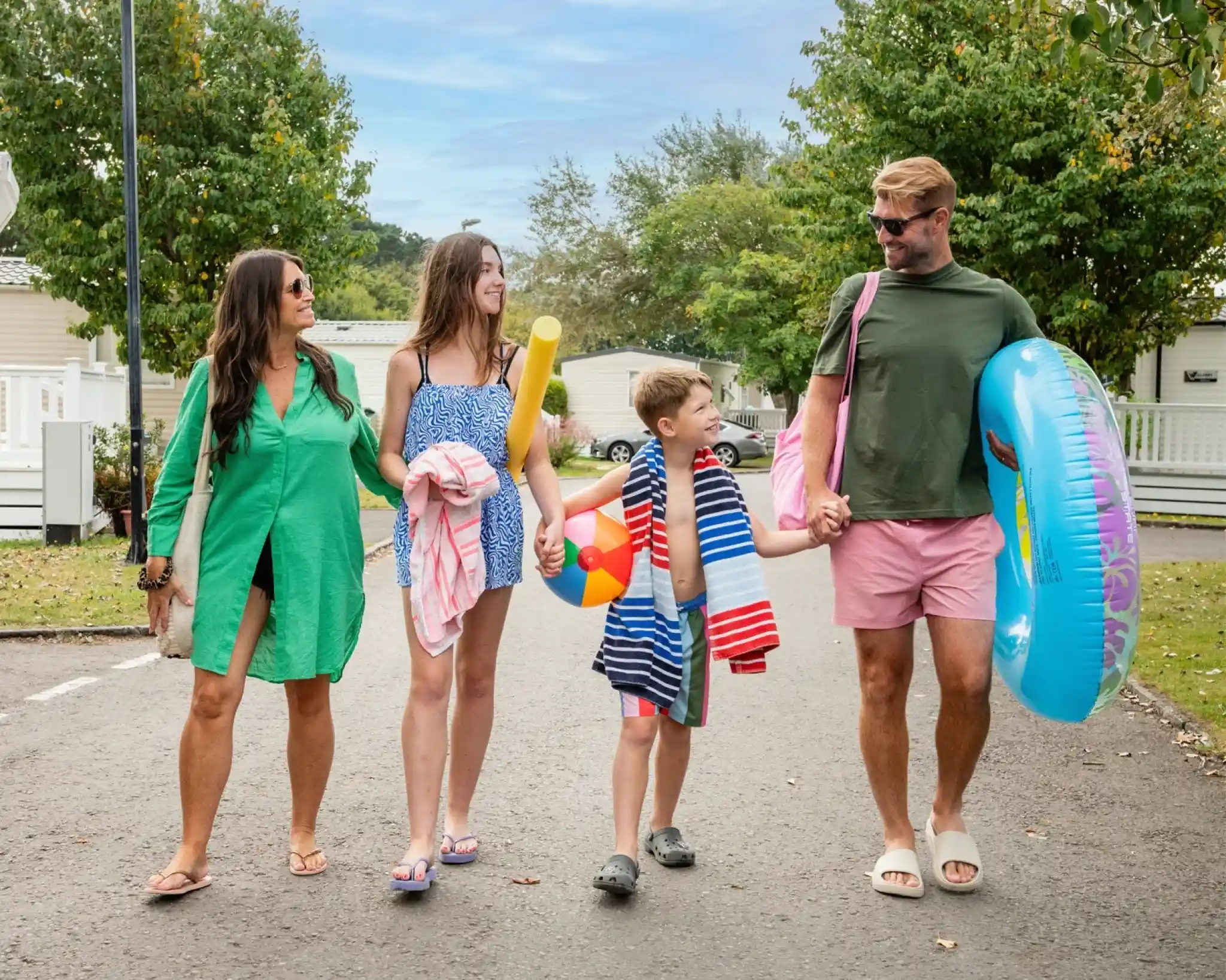 A family walks down a tree-lined road, carrying beach gear. Two adults and two children smile and hold hands, dressed in summer attire. One adult wears a green dress, while the other is in a casual shirt. The children are dressed in swimsuits and hold colorful pool toys.