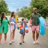 A family walks down a tree-lined road, carrying beach gear. Two adults and two children smile and hold hands, dressed in summer attire. One adult wears a green dress, while the other is in a casual shirt. The children are dressed in swimsuits and hold colorful pool toys.