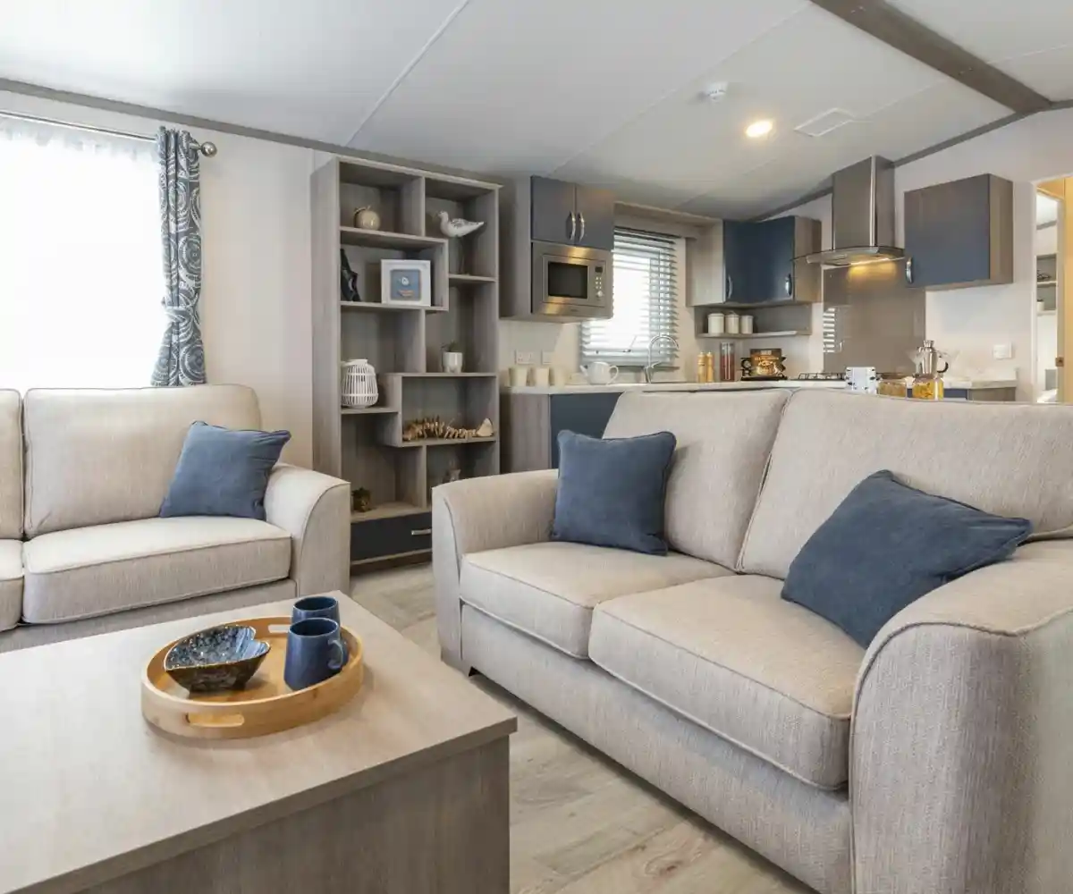 A row of static caravans is set in a lush green area, with a circular flower bed filled with vibrant blooms in the foreground. Tall trees and a clear blue sky complete the tranquil scene.