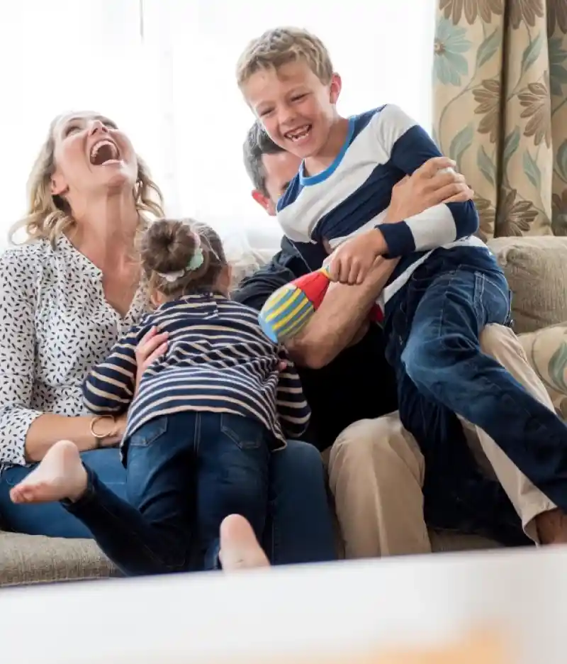 A woman and a man relax on a couch, laughing and enjoying time with a smiling boy who is sitting between them. The boy holds a drink while the woman is looking at him; they are in a bright room with greenery visible outside.
