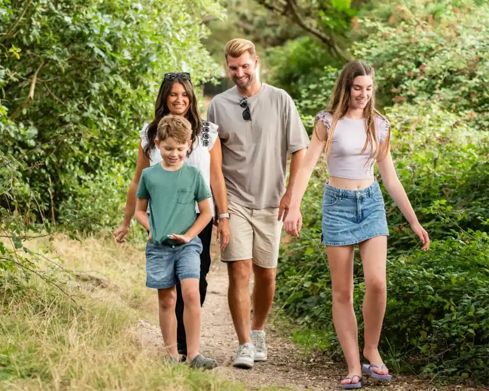 A family walking on a nature trail, smiling and enjoying each other's company. The group includes a woman, a man, a young boy, and a teenage girl, surrounded by greenery and trees.
