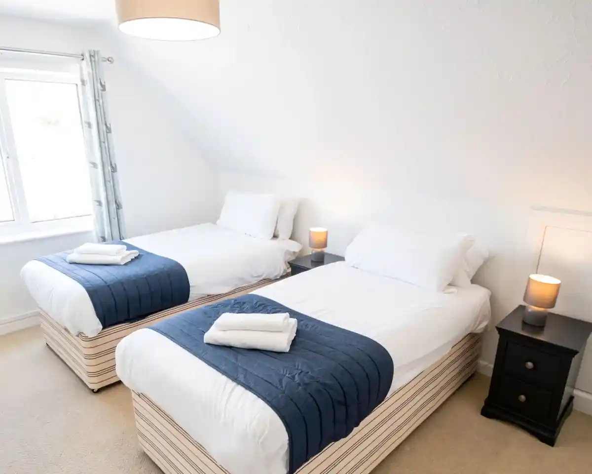 A well-lit bedroom features two single beds with white bedding and navy blue throws. Each bed has a neatly folded towel and a bedside table with a lamp. A window allows natural light to enter the room, enhancing the cozy atmosphere.