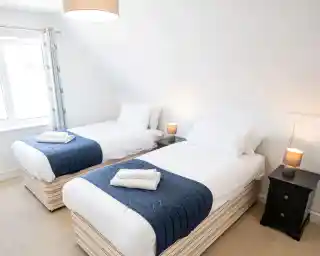 A well-lit bedroom features two single beds with white bedding and navy blue throws. Each bed has a neatly folded towel and a bedside table with a lamp. A window allows natural light to enter the room, enhancing the cozy atmosphere.