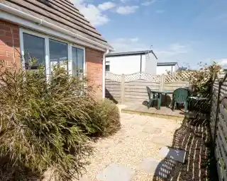 A small outdoor patio area features stone stepping stones leading to a wooden table and chairs surrounded by a gravel garden and shrubbery, under a clear blue sky.