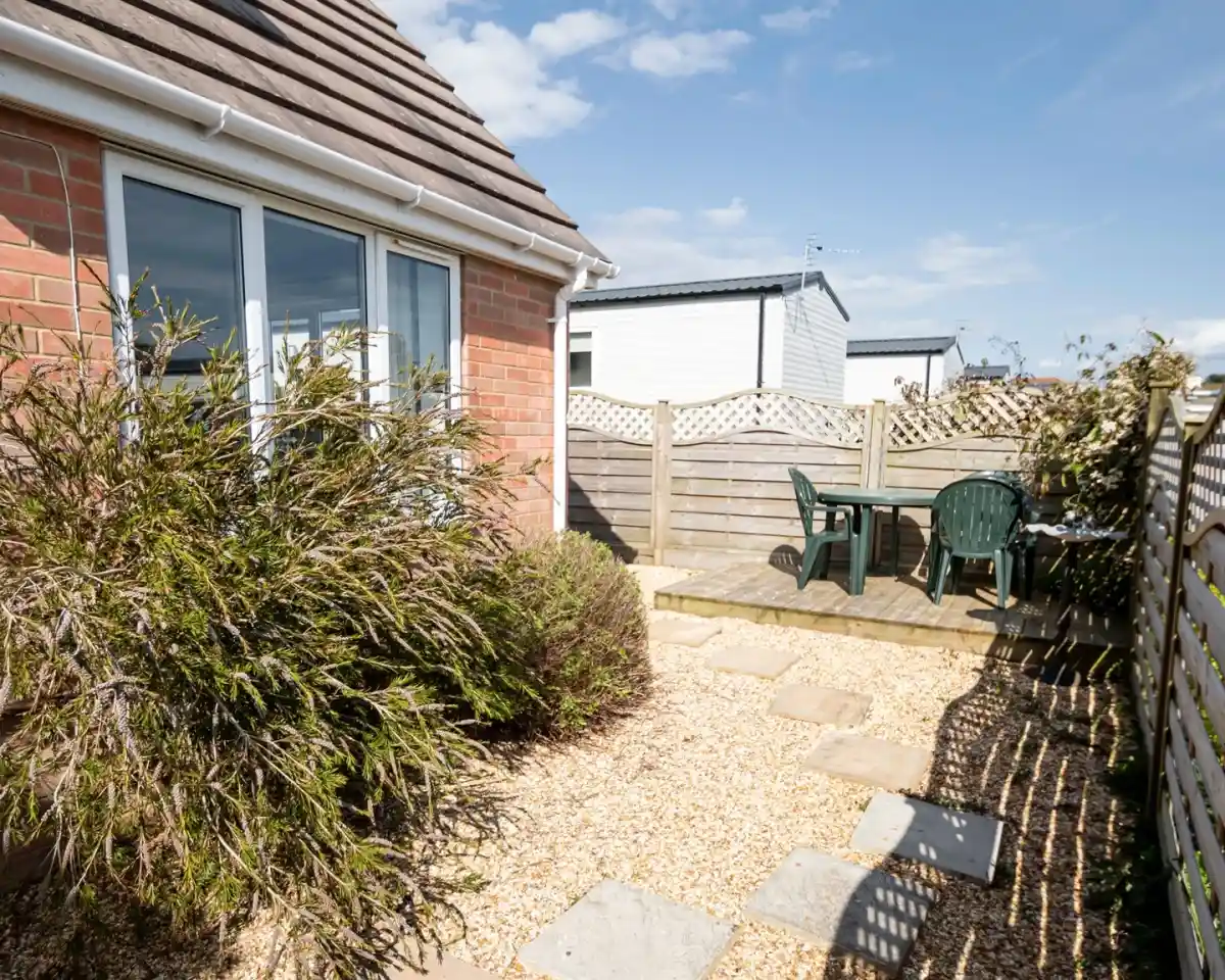 A small outdoor patio area features stone stepping stones leading to a wooden table and chairs surrounded by a gravel garden and shrubbery, under a clear blue sky.