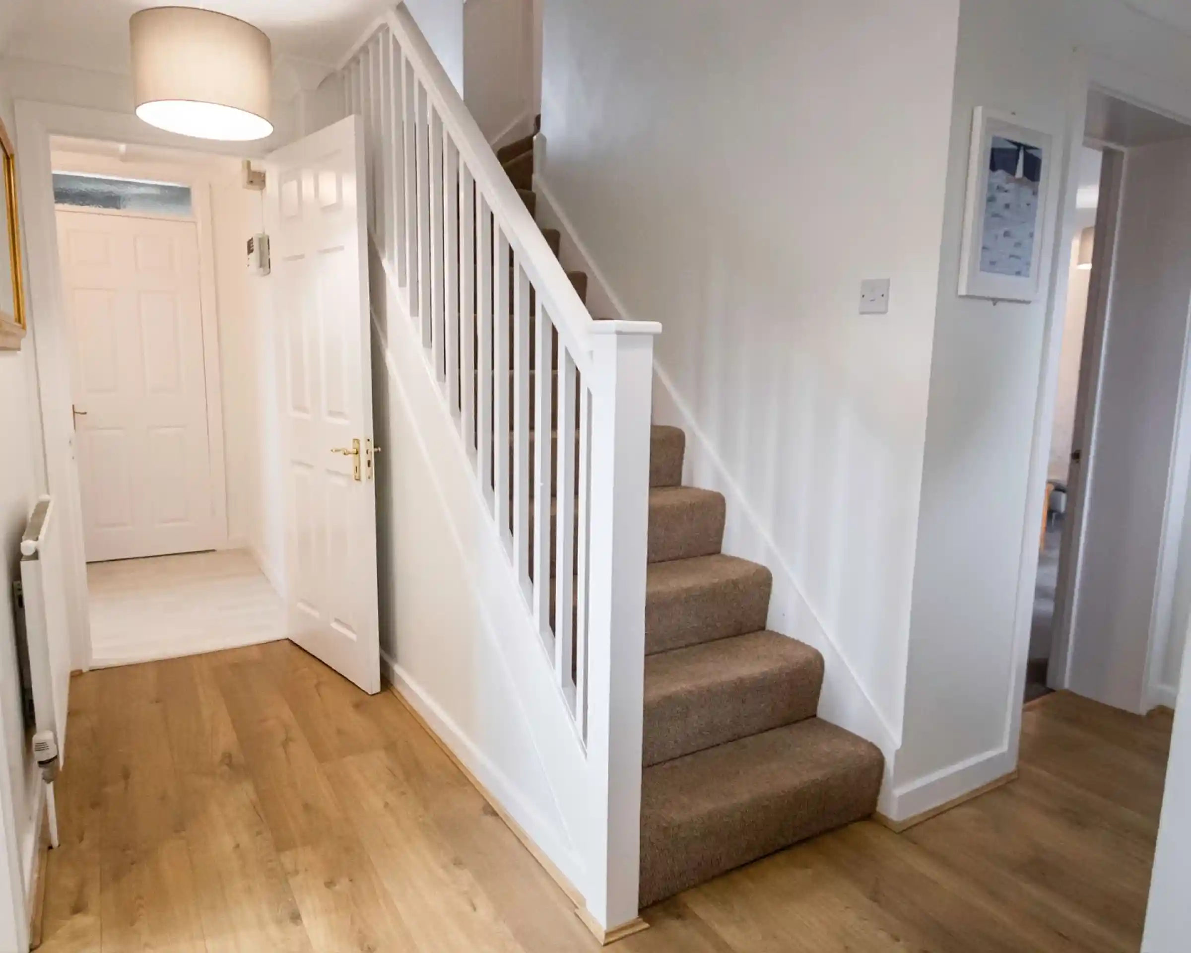 A well-lit hallway featuring a carpeted staircase with white railings. Light-colored walls and wooden flooring create a bright atmosphere. A door is visible on the left, and a small frame is mounted on the wall near the top of the stairs.