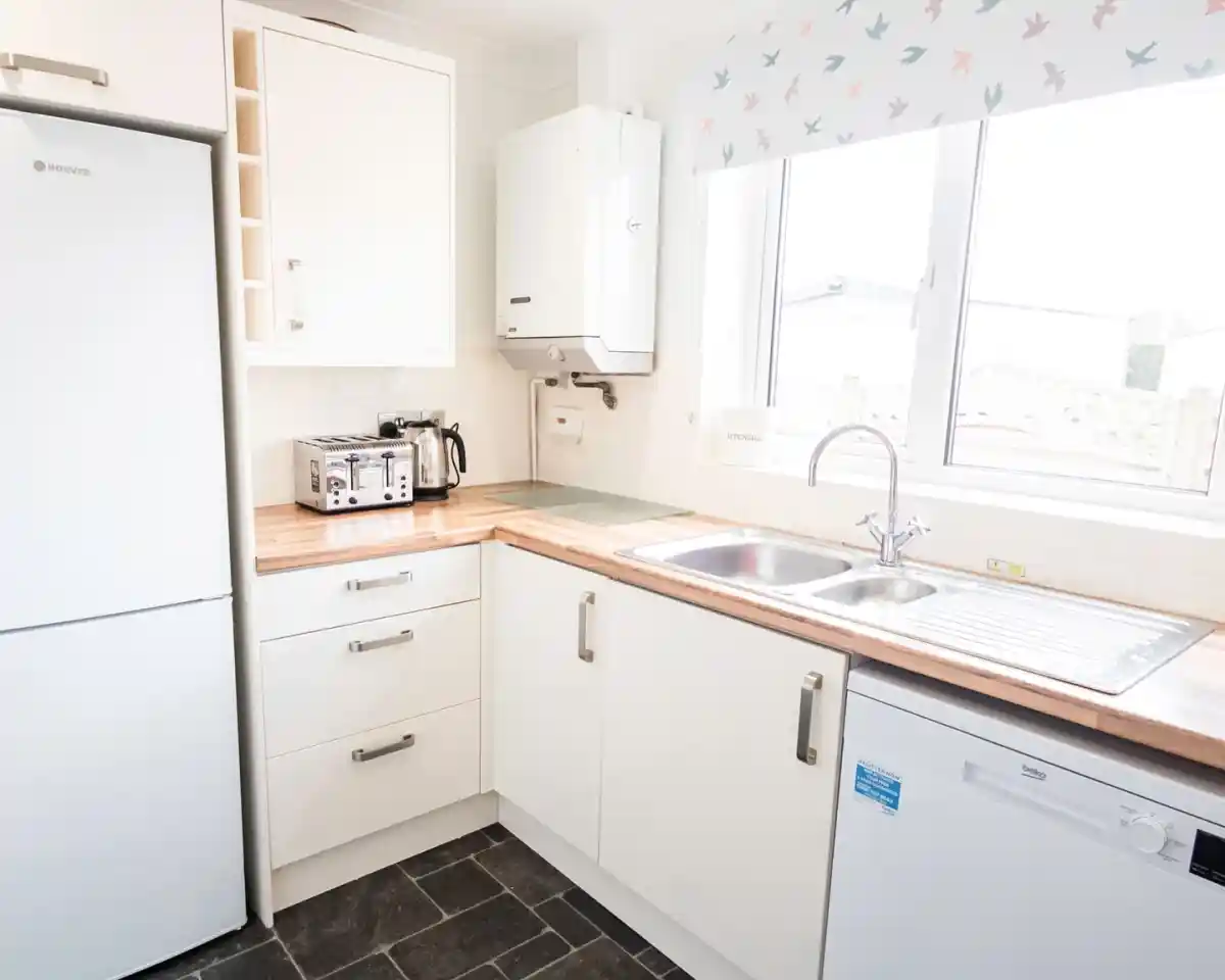 A modern kitchen featuring white cabinetry and a wooden countertop. Stainless steel appliances include a refrigerator and a toaster. A sink with a mixer tap is positioned next to the window, which has a bird-patterned valance. A dishwasher is visible beside the sink.