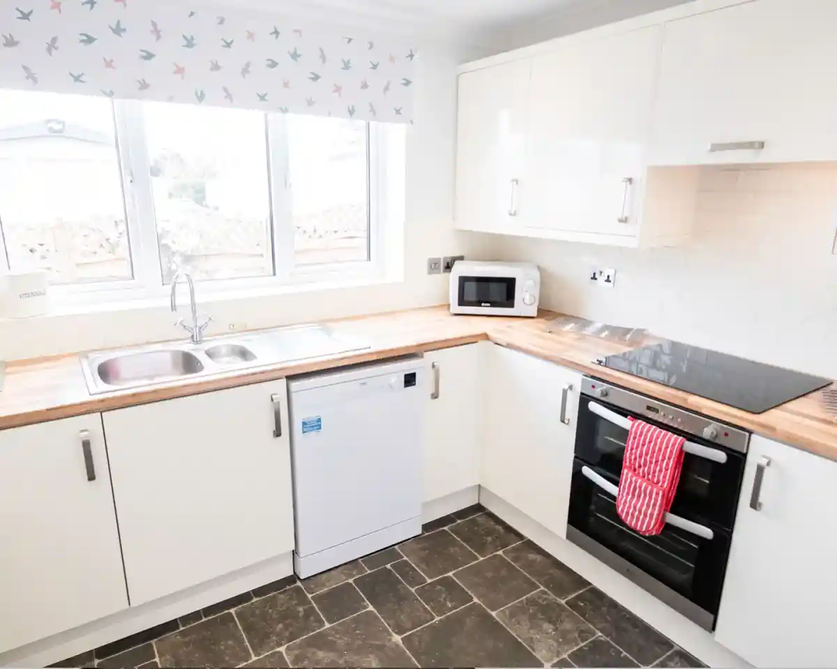 A modern kitchen with white cabinetry and a wooden countertop. It features a double sink, a dishwasher, an electric oven, and a stovetop. A window with a patterned valance overlooks an outdoor area, and the floor has dark tiles.