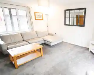 A cozy living room featuring a light gray sectional sofa, a wooden coffee table, and a large window with sheer curtains. A decorative mirror is mounted on the wall, and the floor is covered with soft gray carpet.