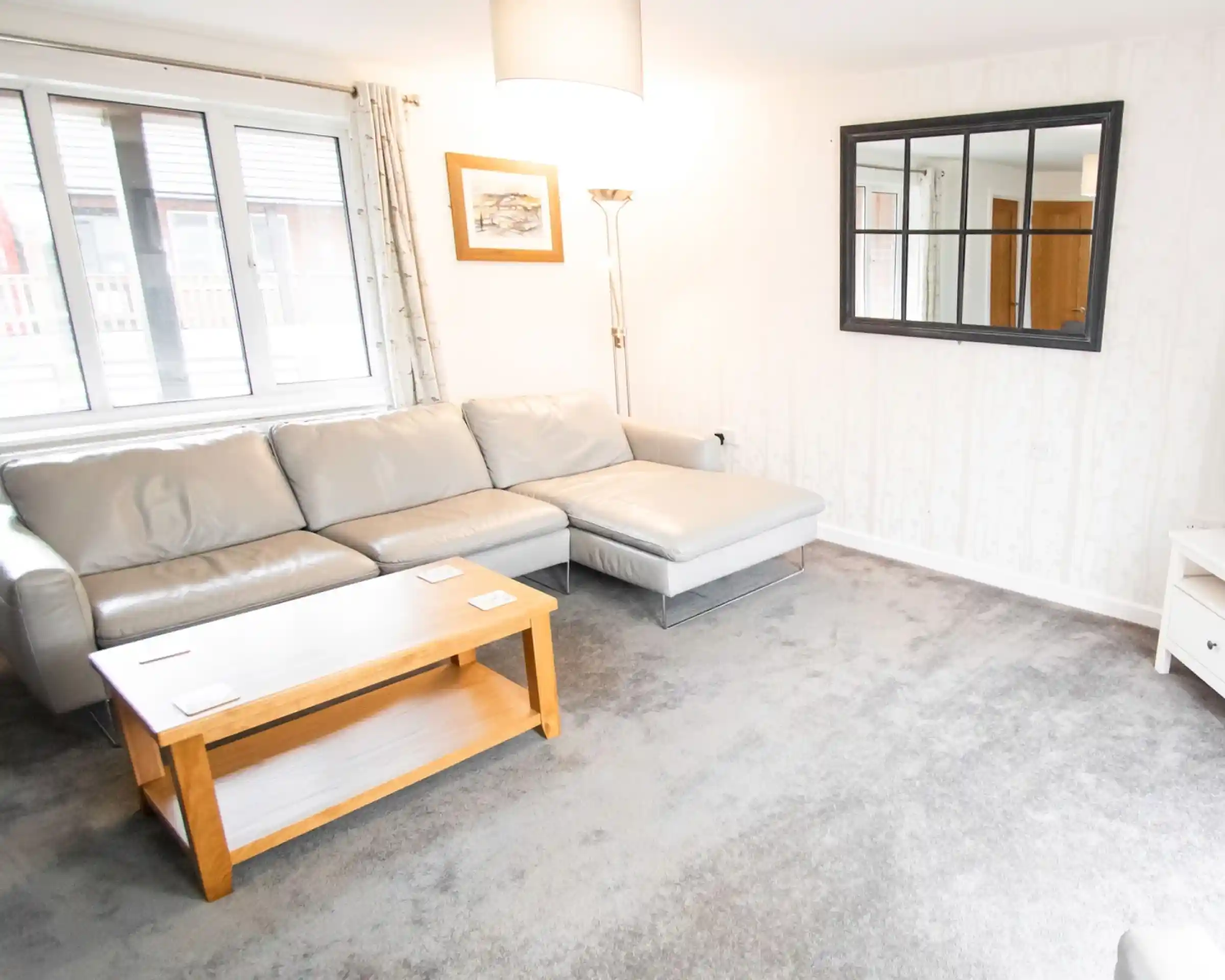 A cozy living room featuring a light gray sectional sofa, a wooden coffee table, and a large window with sheer curtains. A decorative mirror is mounted on the wall, and the floor is covered with soft gray carpet.