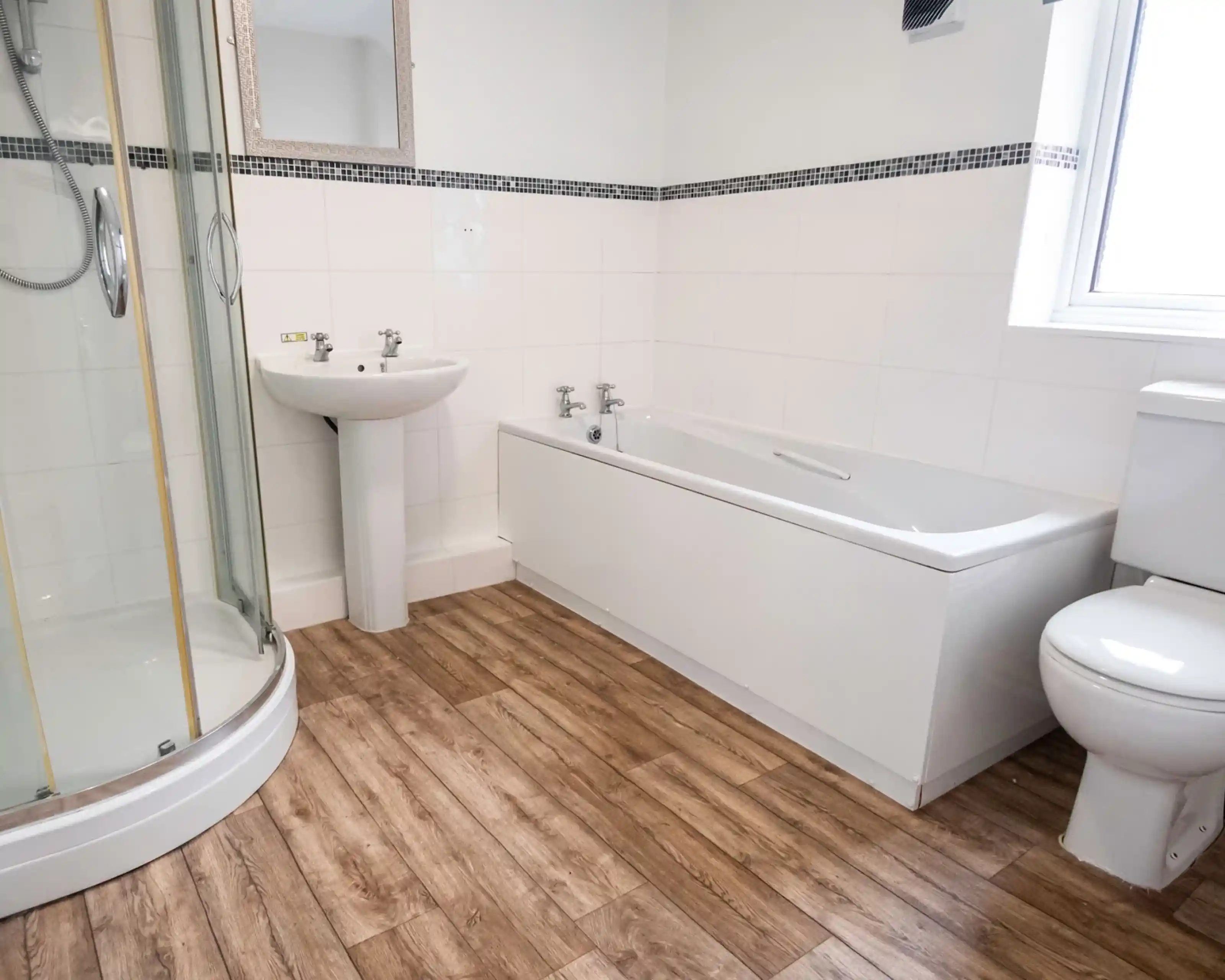 A modern bathroom featuring a glass shower enclosure, bathtub, pedestal sink, and toilet. The flooring is wooden, and the walls are tiled in white with a black accent strip. A mirror is mounted above the sink.