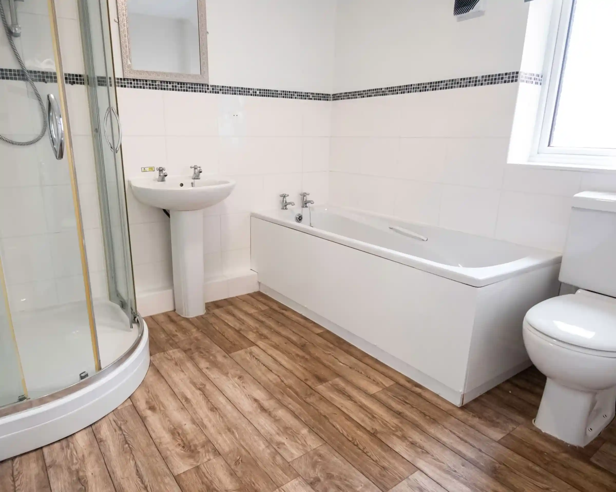 A modern bathroom featuring a glass shower enclosure, bathtub, pedestal sink, and toilet. The flooring is wooden, and the walls are tiled in white with a black accent strip. A mirror is mounted above the sink.