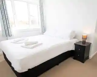 A cozy bedroom featuring a neatly made double bed with white linens and two folded towels on the bed. A bedside table with a lamp sits next to the bed, and large windows allow natural light to brighten the room. Light-colored curtains frame the windows, enhancing the airy atmosphere.