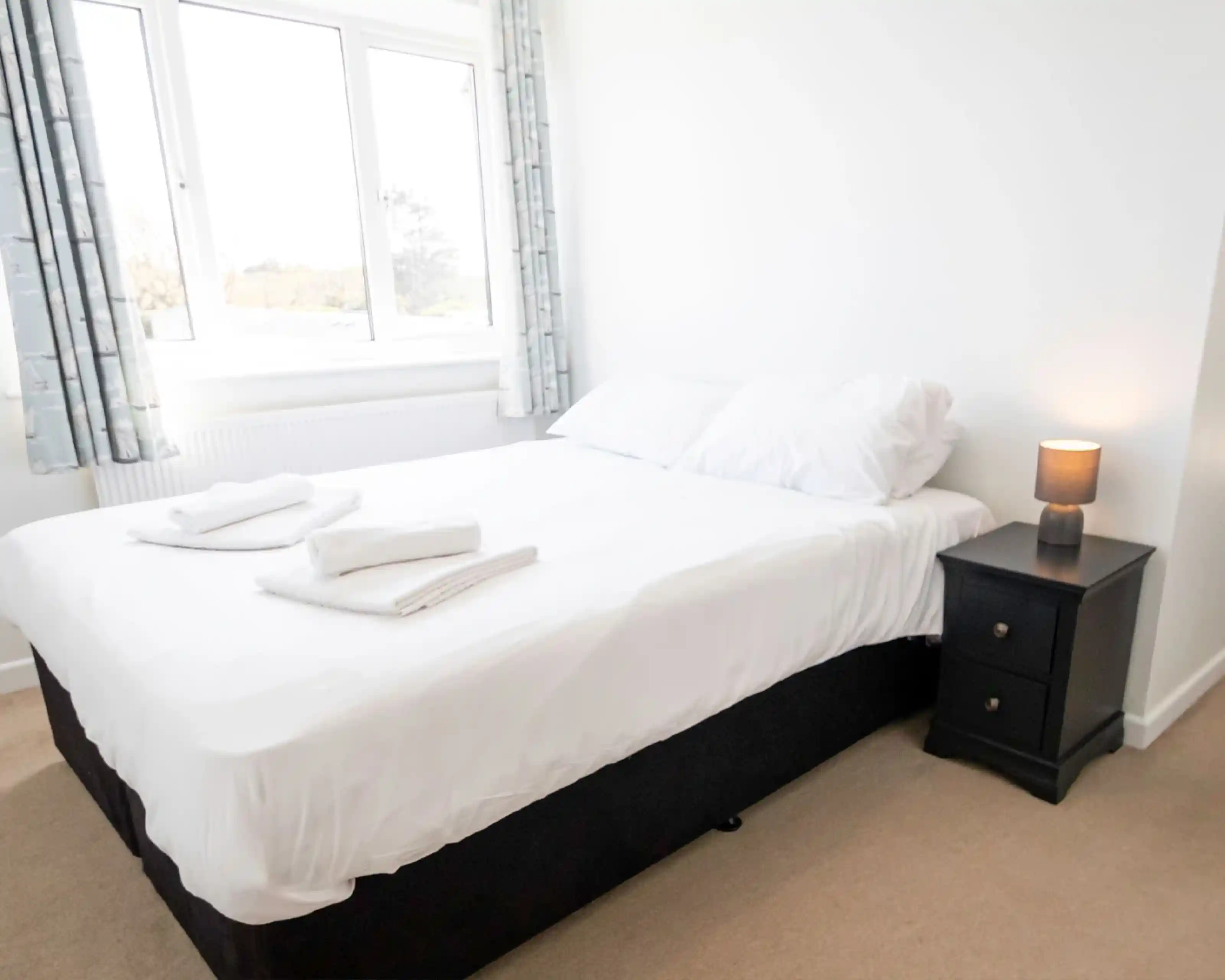 A cozy bedroom featuring a neatly made double bed with white linens and two folded towels on the bed. A bedside table with a lamp sits next to the bed, and large windows allow natural light to brighten the room. Light-colored curtains frame the windows, enhancing the airy atmosphere.