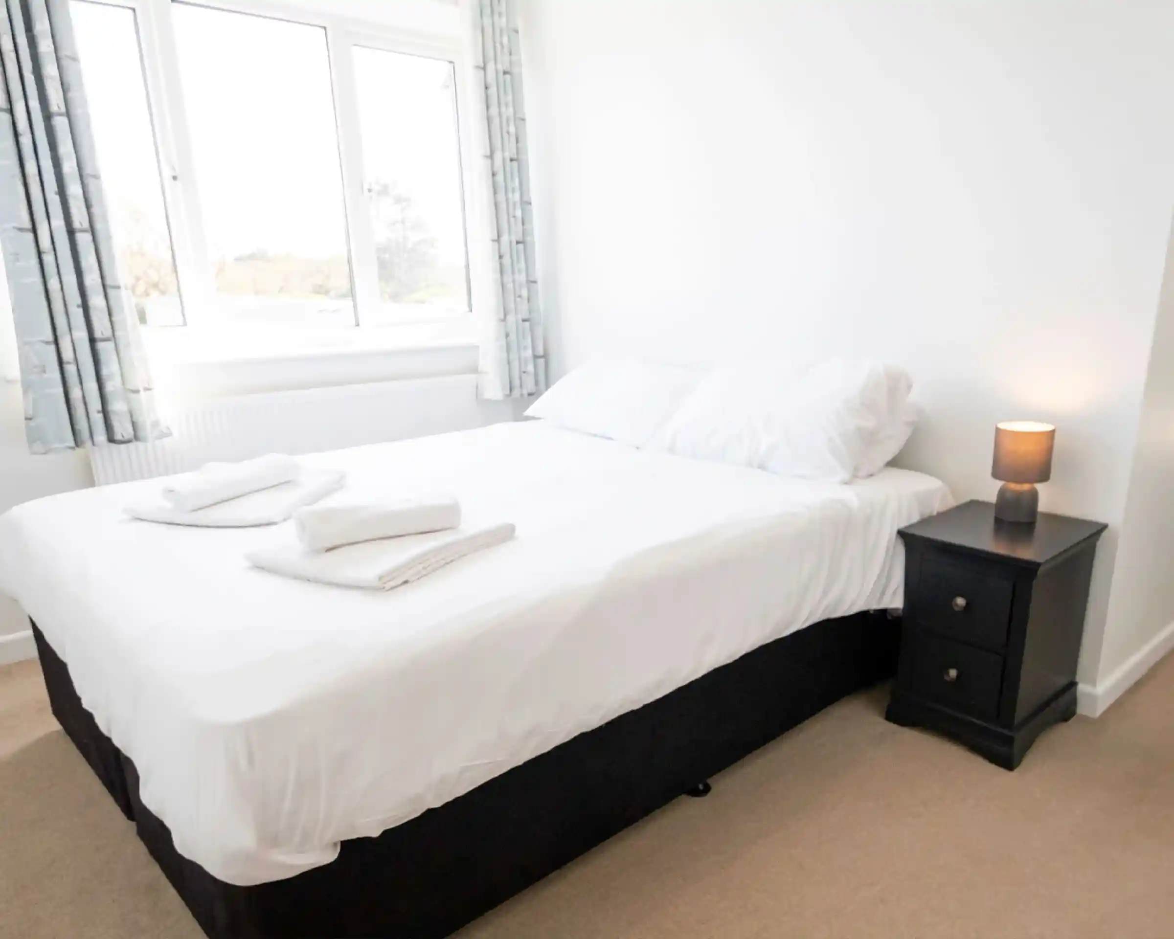 A cozy bedroom featuring a neatly made double bed with white linens and two folded towels on the bed. A bedside table with a lamp sits next to the bed, and large windows allow natural light to brighten the room. Light-colored curtains frame the windows, enhancing the airy atmosphere.