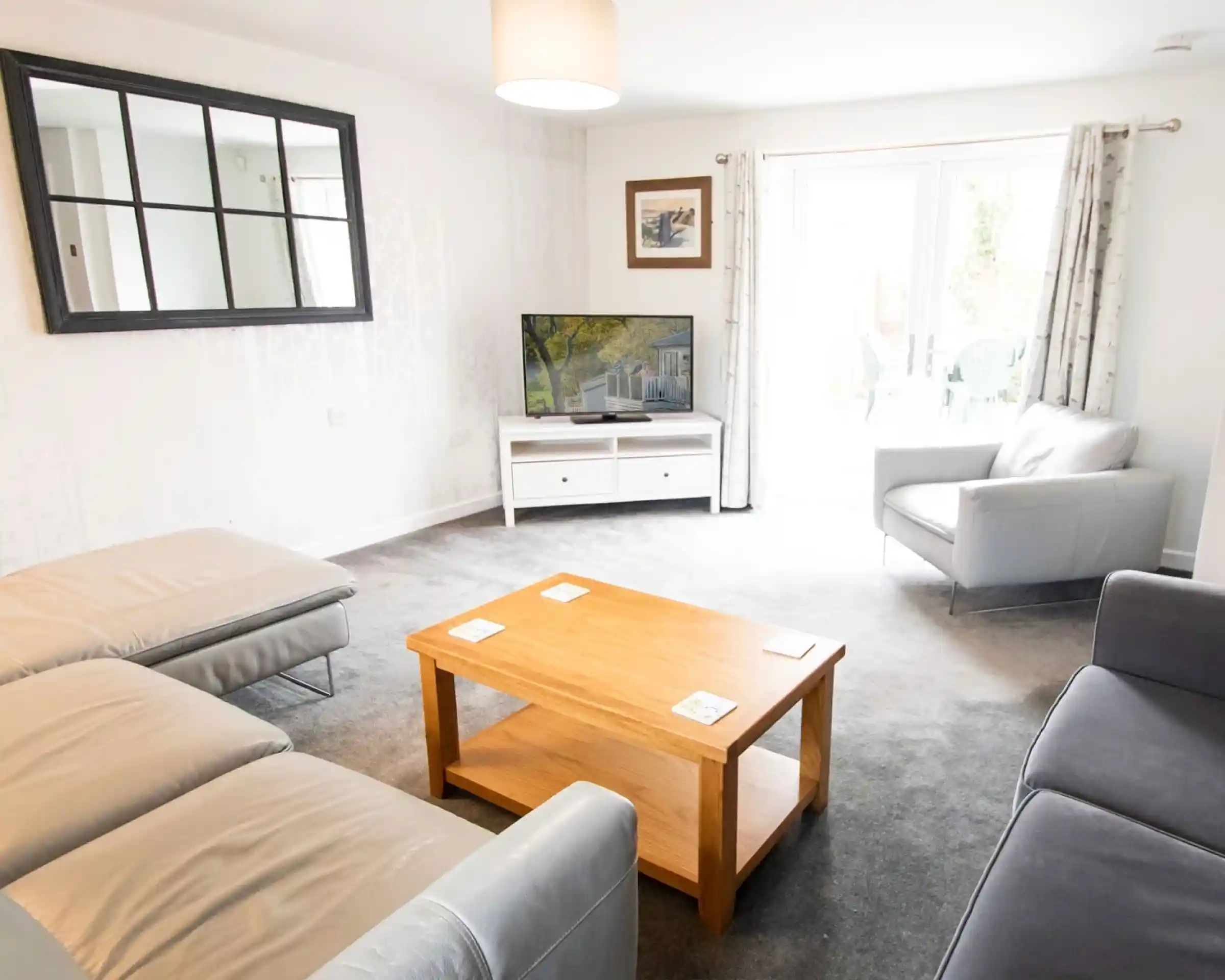 A bright living room featuring a gray sofa and armchair, a wooden coffee table, and a TV on a white cabinet. Natural light streams in through glass doors leading to an outdoor space. A mirror and framed artwork adorn the walls.