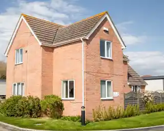 A two-story brick house with a gabled roof, featuring multiple windows and a well-maintained garden. A pathway leads to the entrance, and a fence encloses the yard. The sky is partly cloudy.