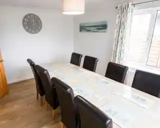A spacious dining area featuring a long wooden table with eight black leather chairs. The table is set with placemats, and there’s a clock on the wall. Natural light streams in through a large window adorned with patterned curtains. A calming piece of art is displayed on the wall.