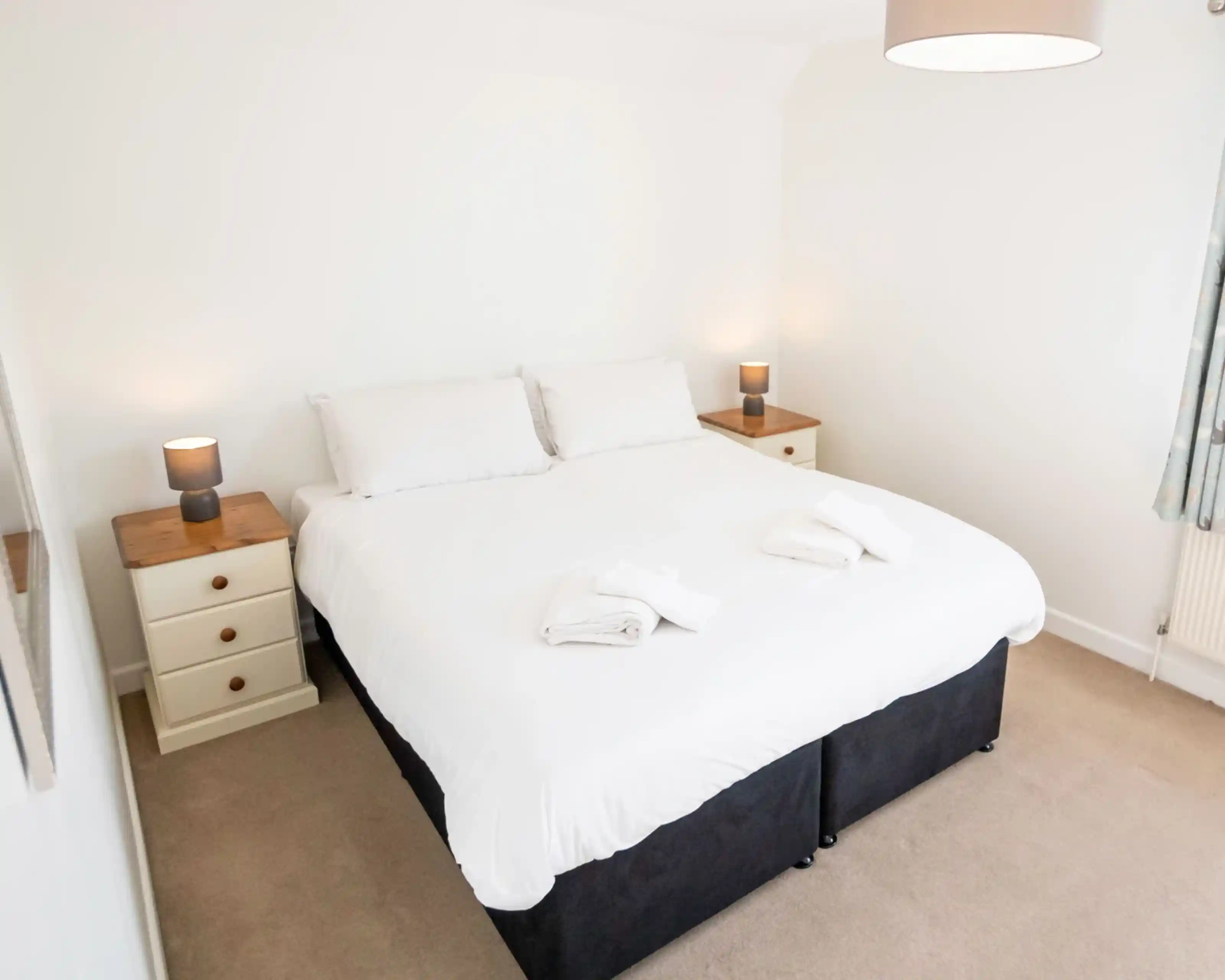 A cozy bedroom featuring a large double bed with white bedding. Two bedside tables flank the bed, each with a small lamp. The walls are painted white, and natural light fills the room. Soft beige carpet covers the floor.