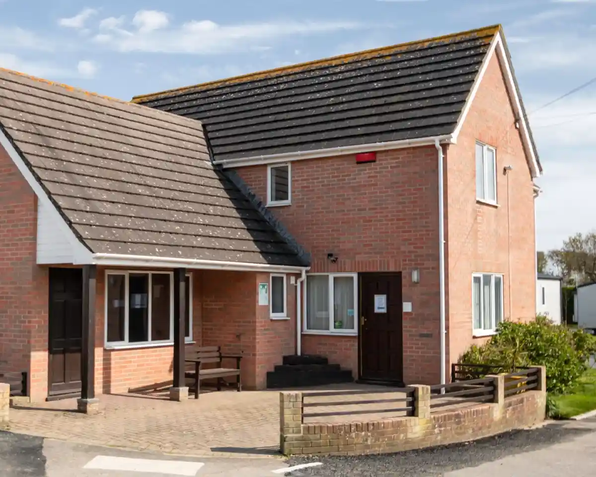 A two-story brick building with a sloped roof, featuring a small porch and a front pathway. The exterior has a mix of red brick and gray shingles, with windows and a door visible. Surrounding the building is a landscaped area with small shrubs.