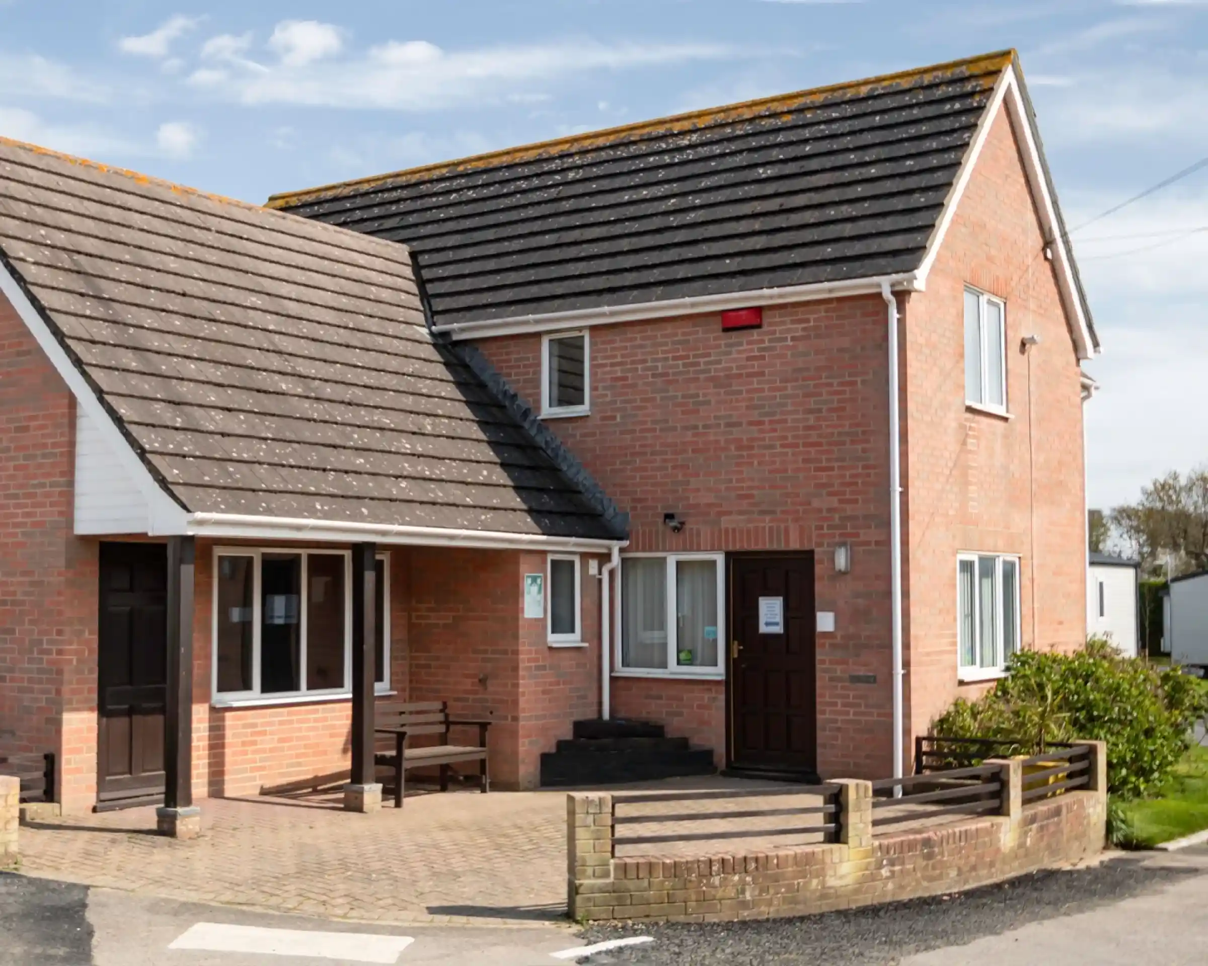 A two-story brick building with a sloped roof, featuring a small porch and a front pathway. The exterior has a mix of red brick and gray shingles, with windows and a door visible. Surrounding the building is a landscaped area with small shrubs.
