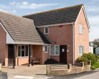 A two-story brick building with a sloped roof, featuring a small porch and a front pathway. The exterior has a mix of red brick and gray shingles, with windows and a door visible. Surrounding the building is a landscaped area with small shrubs.