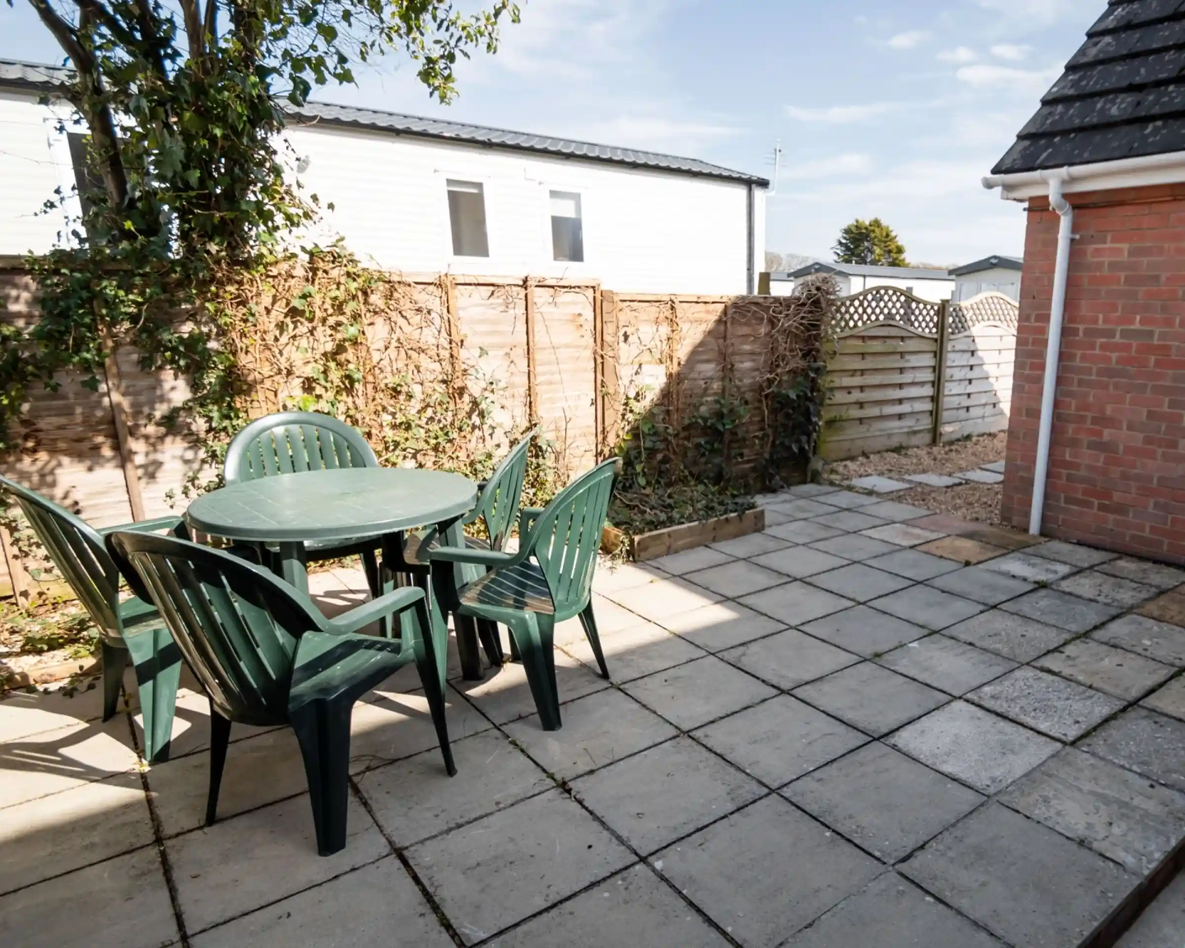 An outdoor patio area featuring a round green table surrounded by four green chairs. The space is bordered by wooden fencing, with plants climbing the fence and a gravel area to one side. A bright sky is visible above.