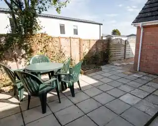 An outdoor patio area featuring a round green table surrounded by four green chairs. The space is bordered by wooden fencing, with plants climbing the fence and a gravel area to one side. A bright sky is visible above.