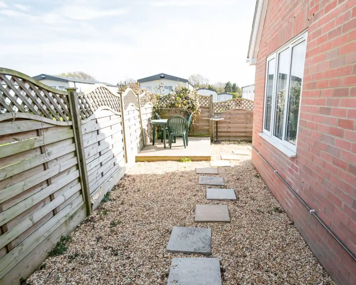 A narrow pathway lined with stone slabs leads to a wooden patio area with a table and chairs, surrounded by wooden fences and flowering plants. A clear sky is visible in the background.