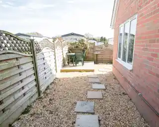 A narrow pathway lined with stone slabs leads to a wooden patio area with a table and chairs, surrounded by wooden fences and flowering plants. A clear sky is visible in the background.