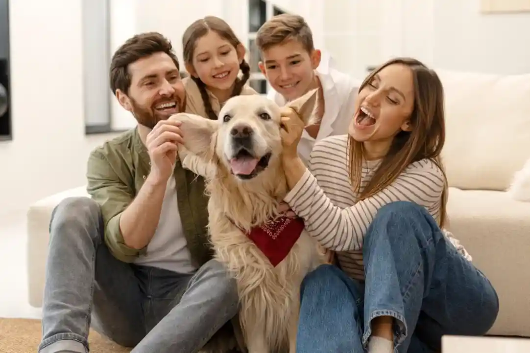 A family of four sits on a cozy living room floor, smiling and playing with a golden retriever wearing a red bandana. The parents and two children are enjoying a fun moment together, with playful expressions and laughter.