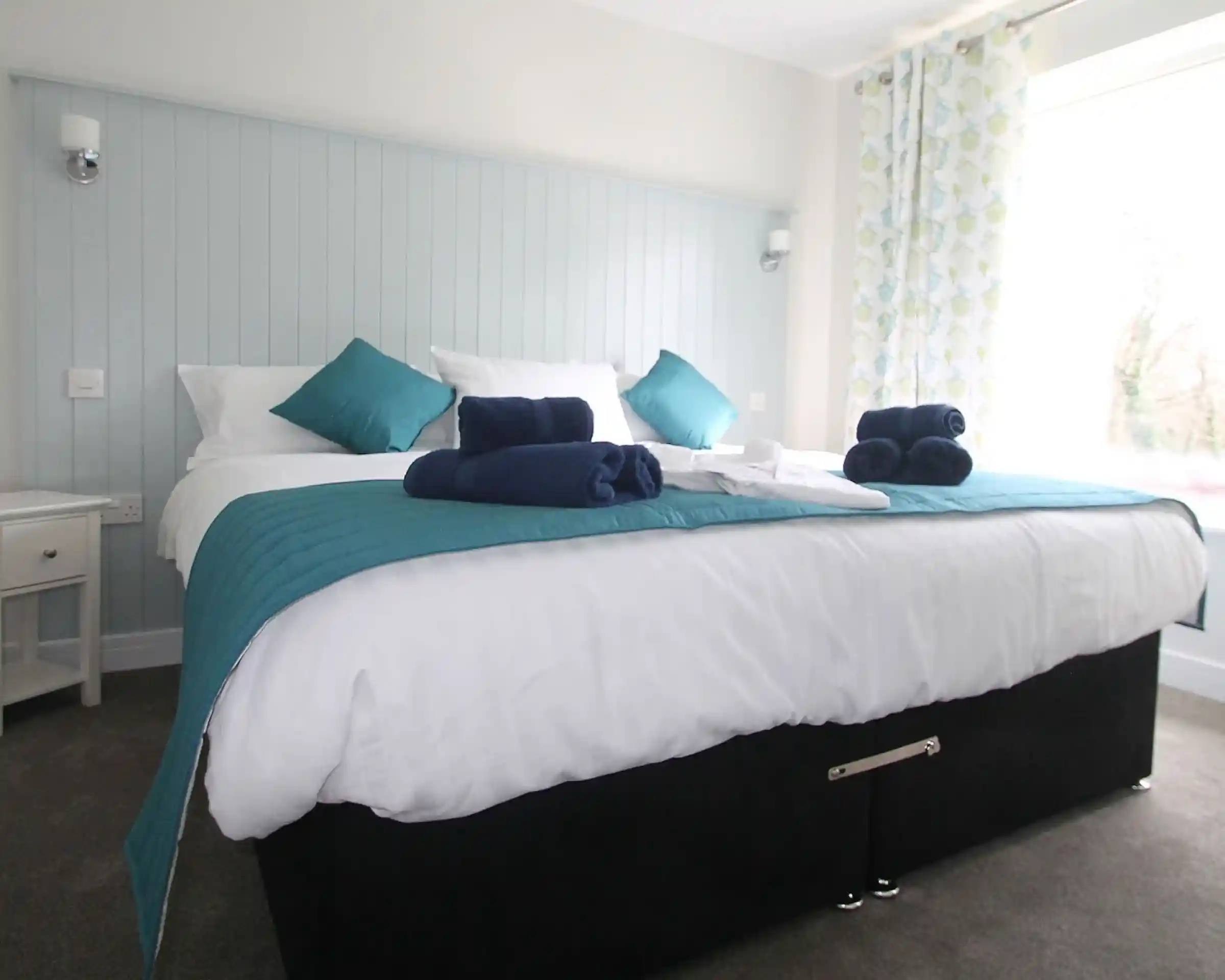 A neatly made bed with a turquoise and white color scheme, featuring plush pillows and rolled towels on the bed. Soft curtains with a floral pattern are next to a large window, allowing natural light into the room.