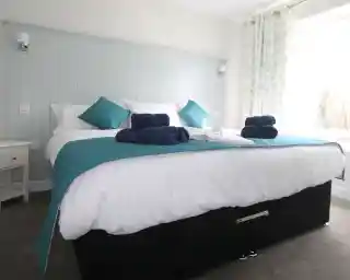 A neatly made bed with a turquoise and white color scheme, featuring plush pillows and rolled towels on the bed. Soft curtains with a floral pattern are next to a large window, allowing natural light into the room.