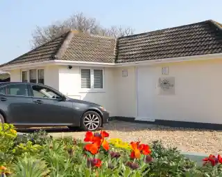 A modern bungalow with a grey car parked in front and colorful flowers in the foreground. The house features a textured roof and an inviting sign beside the entrance.