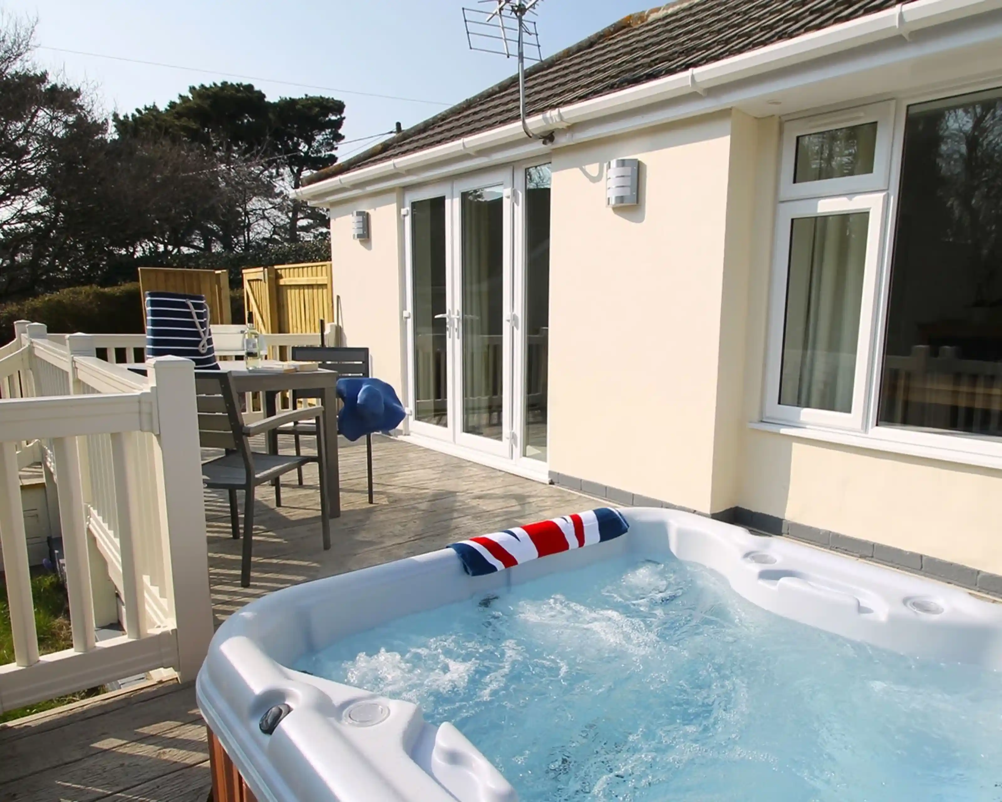 A hot tub with a British flag design is on a wooden deck beside a modern house. Nearby, there are outdoor chairs and a table, with sliding glass doors leading indoors. Trees are visible in the background under a clear sky.
