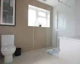 Modern bathroom featuring a glass walk-in shower, a white toilet, and a minimalist sink area. Natural light enters through a window, and a small plant is placed on the sill. The walls are adorned with a subtle textured design, and a framed artwork is visible.