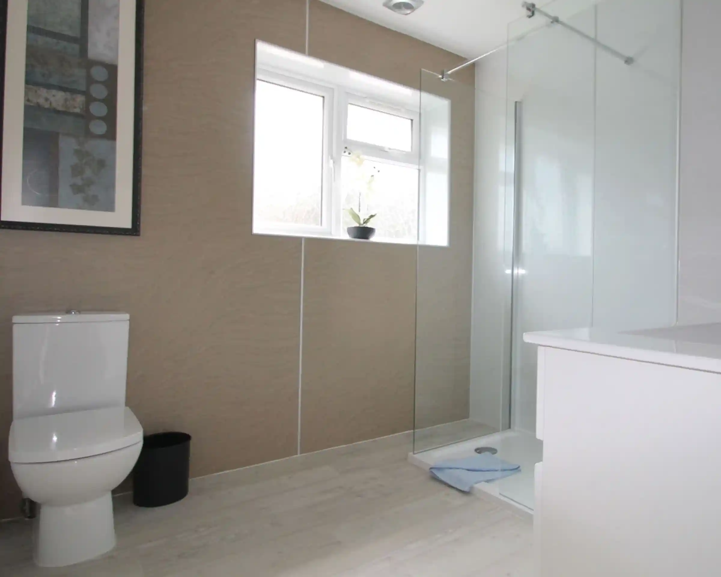 Modern bathroom featuring a glass walk-in shower, a white toilet, and a minimalist sink area. Natural light enters through a window, and a small plant is placed on the sill. The walls are adorned with a subtle textured design, and a framed artwork is visible.