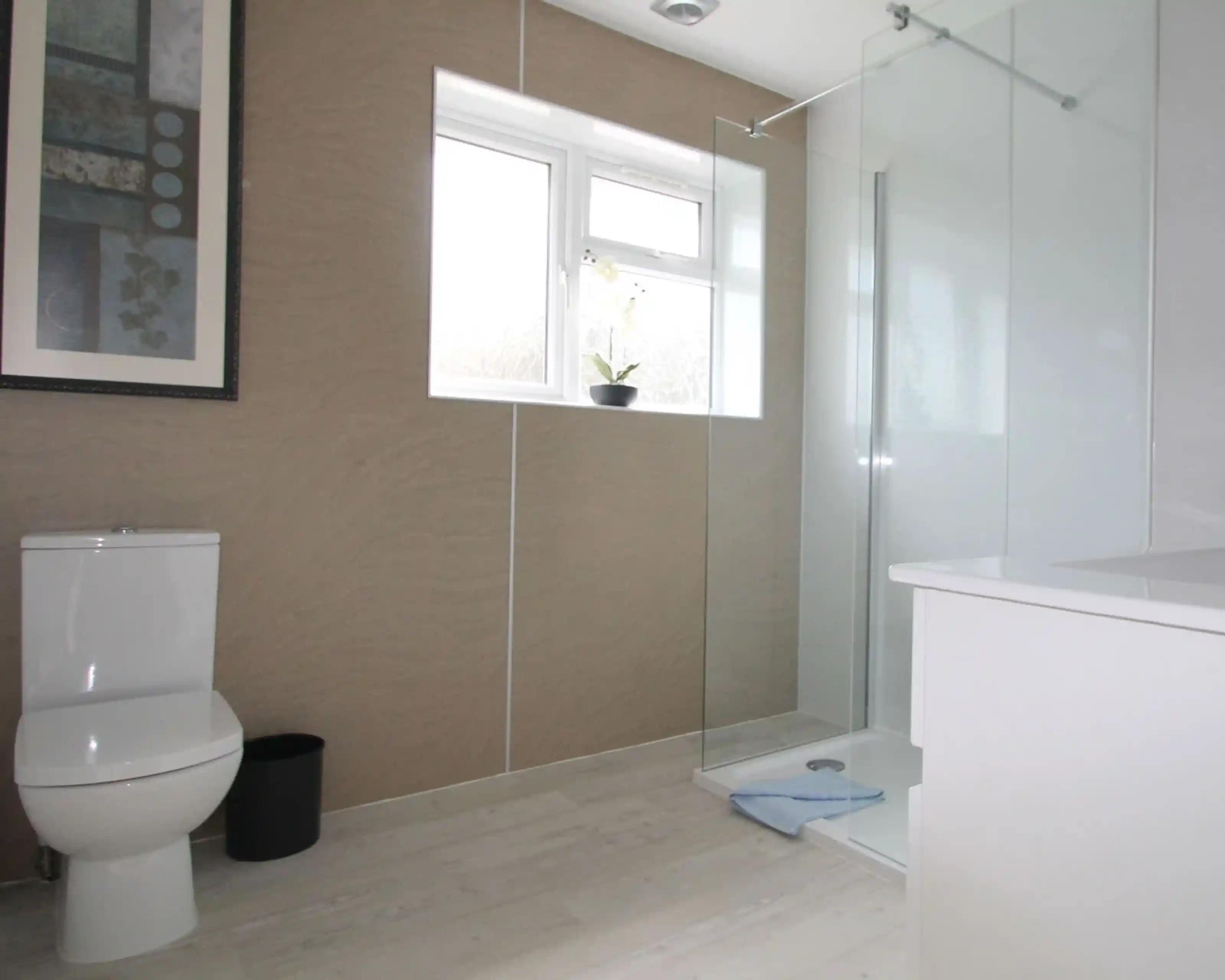 Modern bathroom featuring a glass walk-in shower, a white toilet, and a minimalist sink area. Natural light enters through a window, and a small plant is placed on the sill. The walls are adorned with a subtle textured design, and a framed artwork is visible.