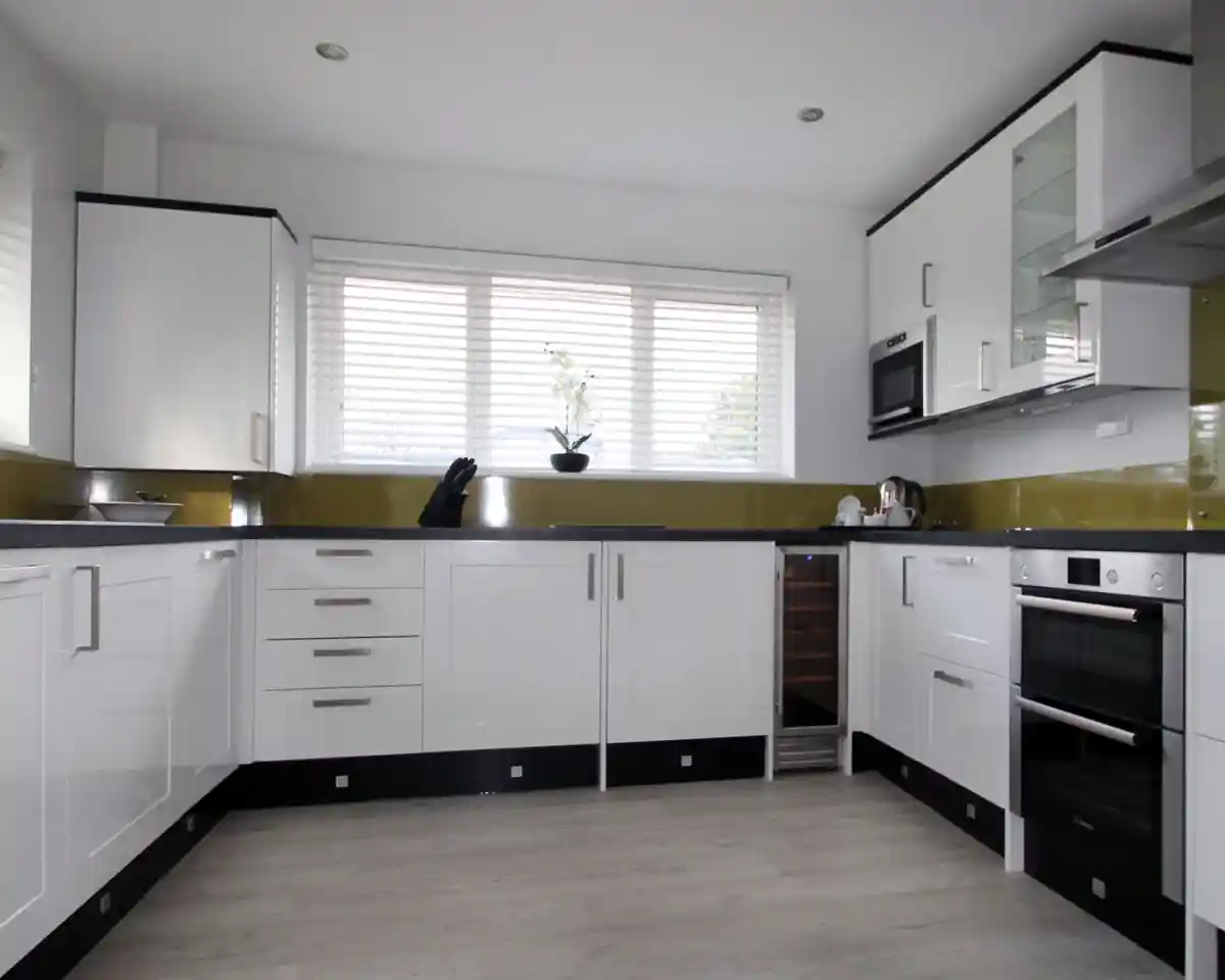 Modern kitchen featuring sleek white cabinets, stainless steel appliances, and a green backsplash. Natural light filters through large windows adorned with blinds. A minimalist design is emphasized with a wooden floor and organized countertop.