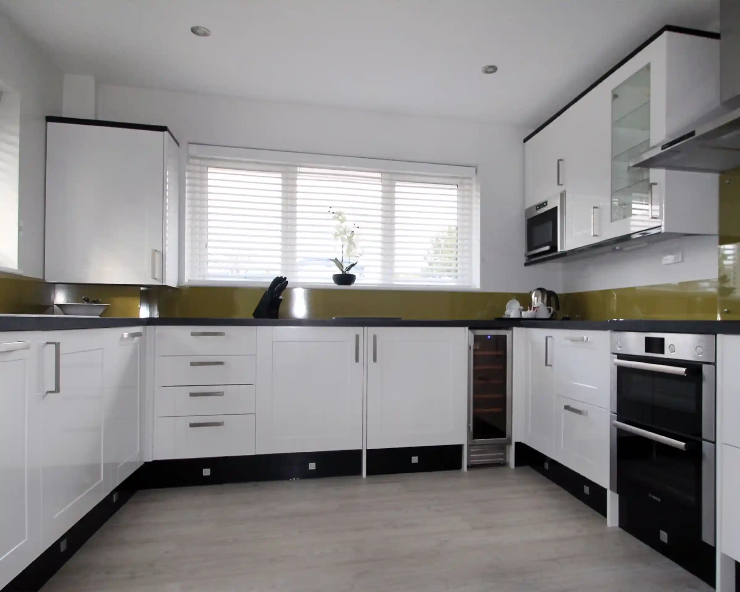 Modern kitchen featuring sleek white cabinets, stainless steel appliances, and a green backsplash. Natural light filters through large windows adorned with blinds. A minimalist design is emphasized with a wooden floor and organized countertop.