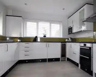 Modern kitchen featuring sleek white cabinets, stainless steel appliances, and a green backsplash. Natural light filters through large windows adorned with blinds. A minimalist design is emphasized with a wooden floor and organized countertop.