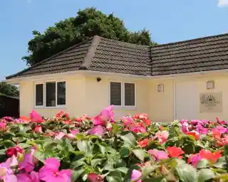 Bright yellow cottage with a sloped roof surrounded by vibrant pink and green flowers, against a clear blue sky.