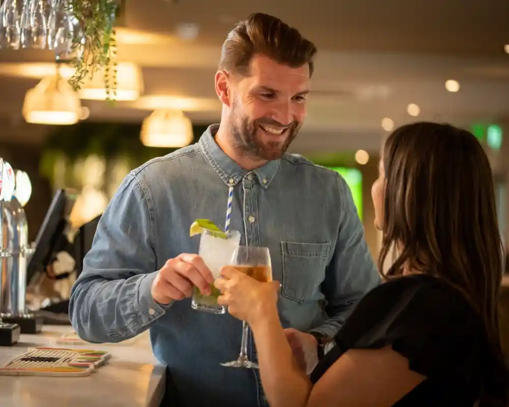 A man with a beard smiles while handing a cocktail to a woman at a bar. The woman, wearing a black top, holds a drink and appears engaged in conversation. The setting is warm and inviting, with soft lighting and greenery in the background.