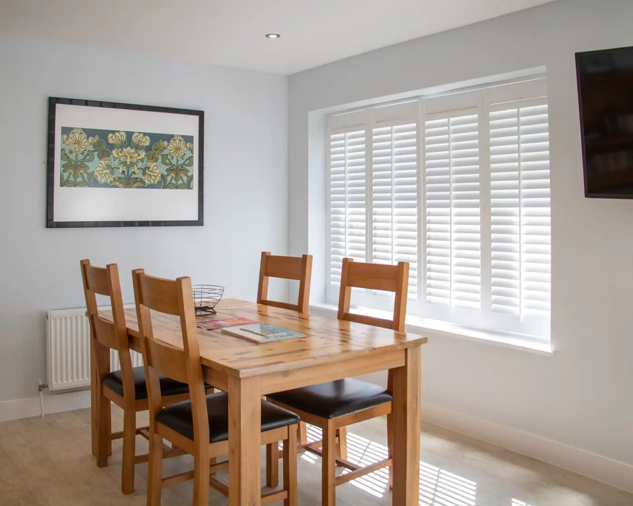 A light-filled dining area features a wooden table surrounded by six chairs. A modern artwork hangs on the wall above the table, and elegant white shutters cover the window. A television is mounted on the opposite wall.