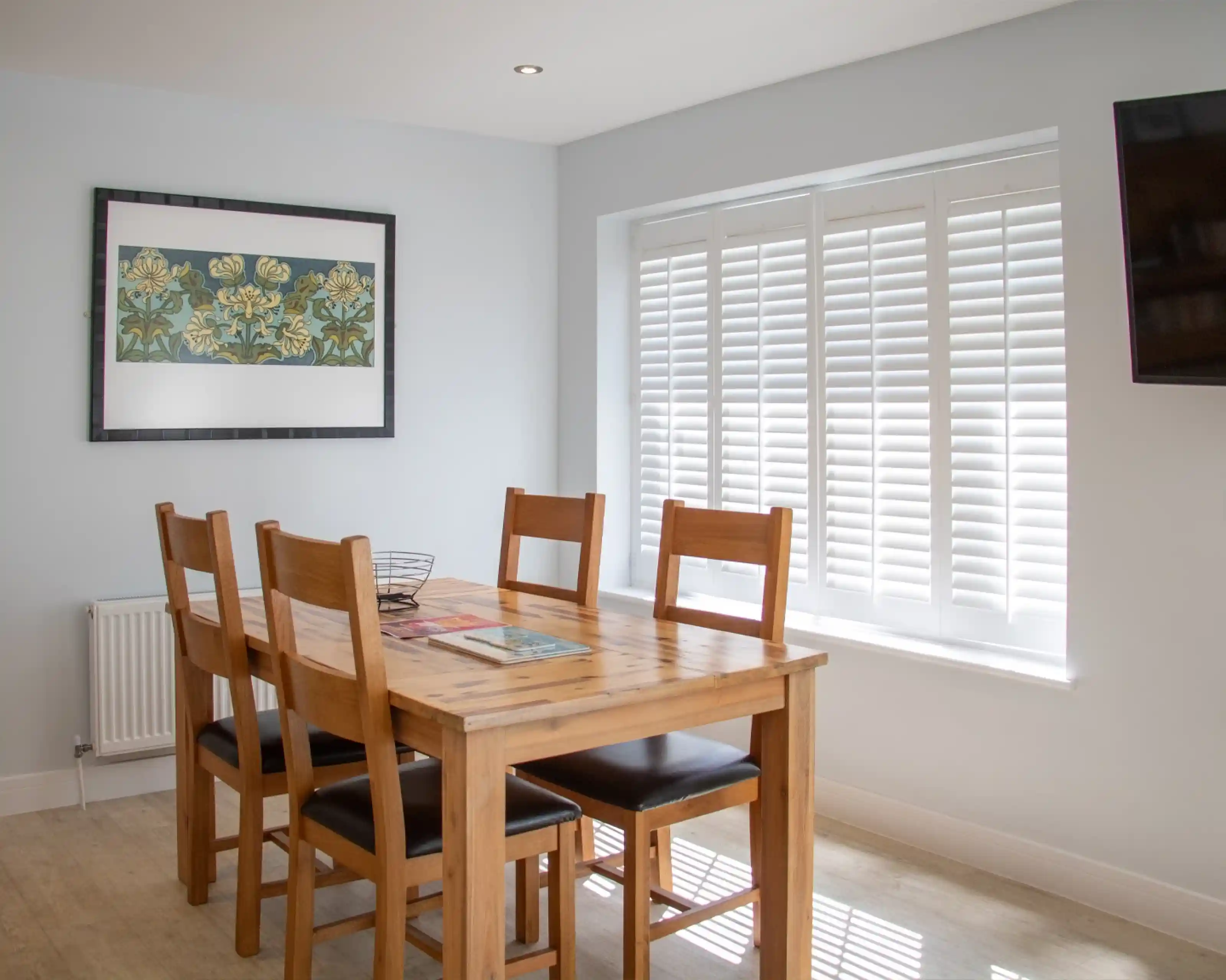 A light-filled dining area features a wooden table surrounded by six chairs. A modern artwork hangs on the wall above the table, and elegant white shutters cover the window. A television is mounted on the opposite wall.