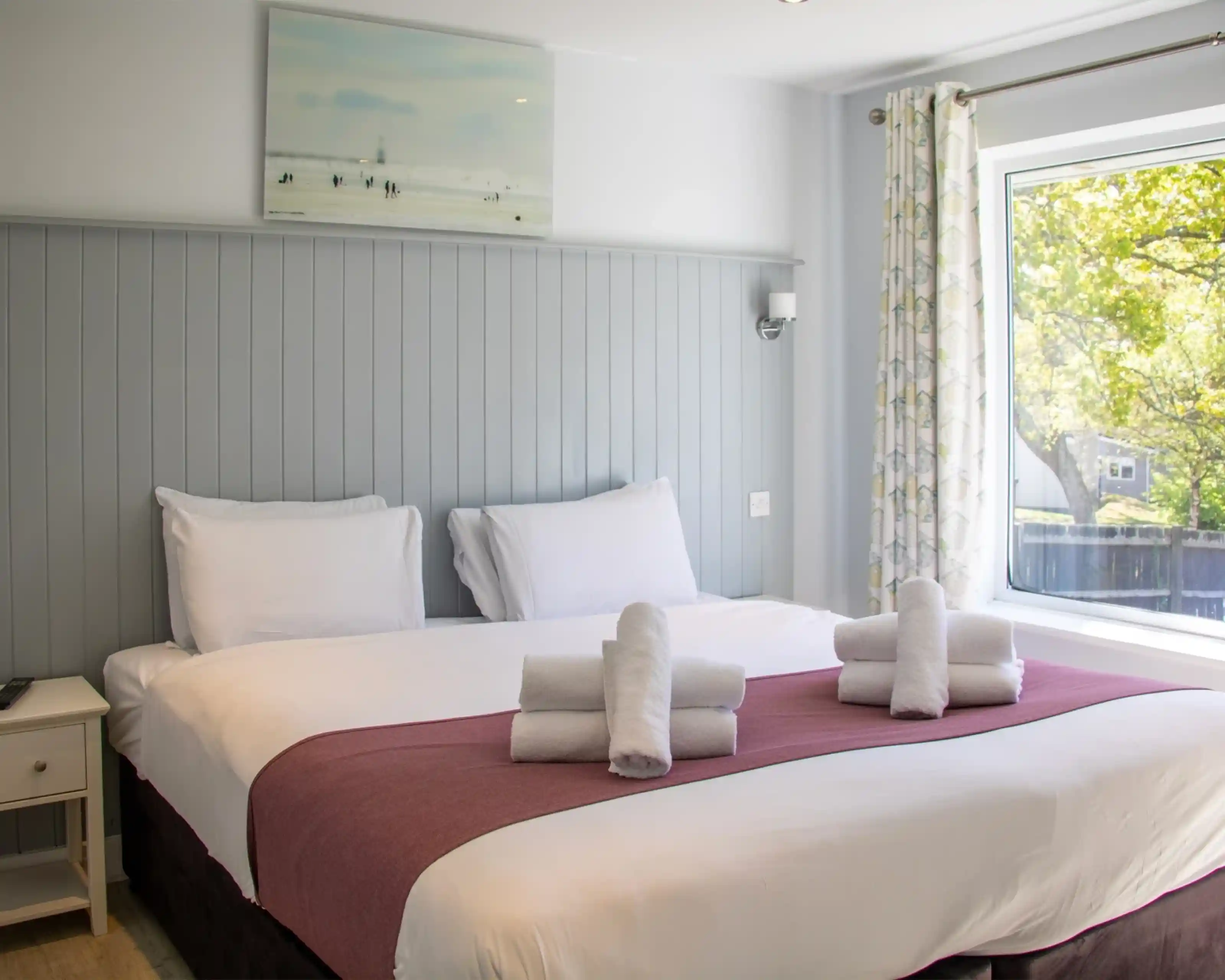 A cozy hotel room featuring a large bed with white linens and a purple accent. Towels are neatly arranged on the bed, and a large window provides natural light, showcasing green trees outside. The walls are painted light gray, and there's a modern artwork above the bed.