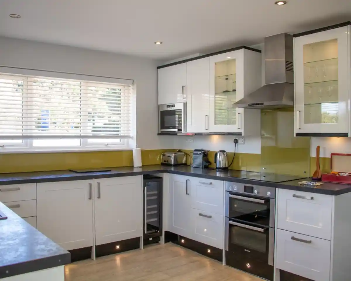 Modern kitchen featuring white and black cabinetry, a stainless steel oven, a built-in microwave, and a wine cooler. There are countertop appliances like a toaster and kettle, with natural light streaming through a window with blinds. A sleek design and clean lines are emphasized throughout the space.