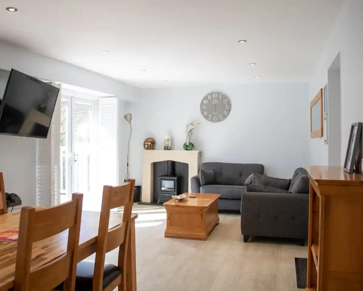 Cozy living room featuring a gray sofa, wooden coffee table, and a fireplace. A large wall clock and television are visible, along with a dining area set with wooden chairs. Natural light streams in from sliding doors.