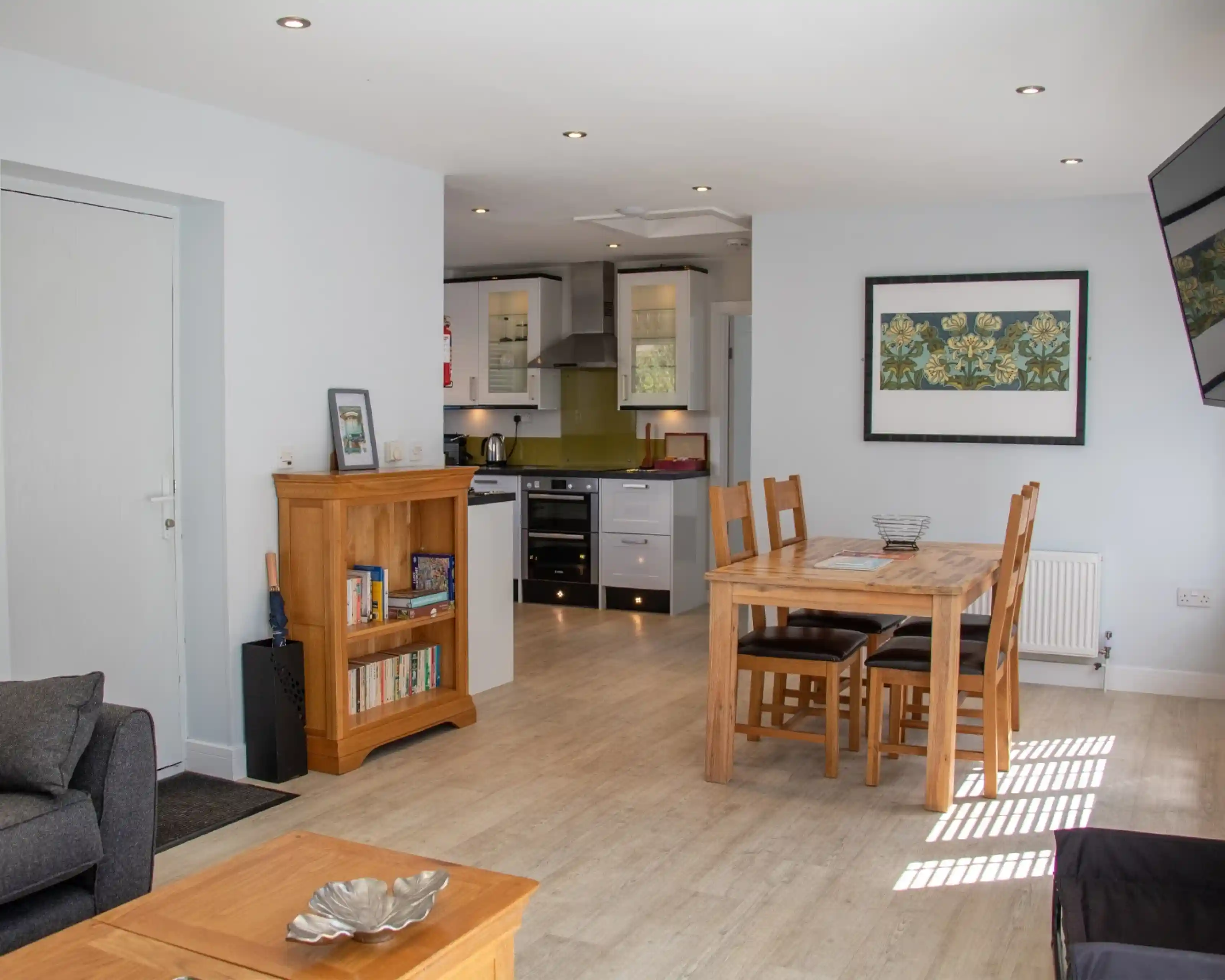 A modern, open-concept living space featuring a cozy seating area with a dark sofa, a wooden dining table with four chairs, and a kitchen area in the background. The kitchen has white cabinets and a vibrant green backsplash. Natural light streams in through a window, illuminating the room.
